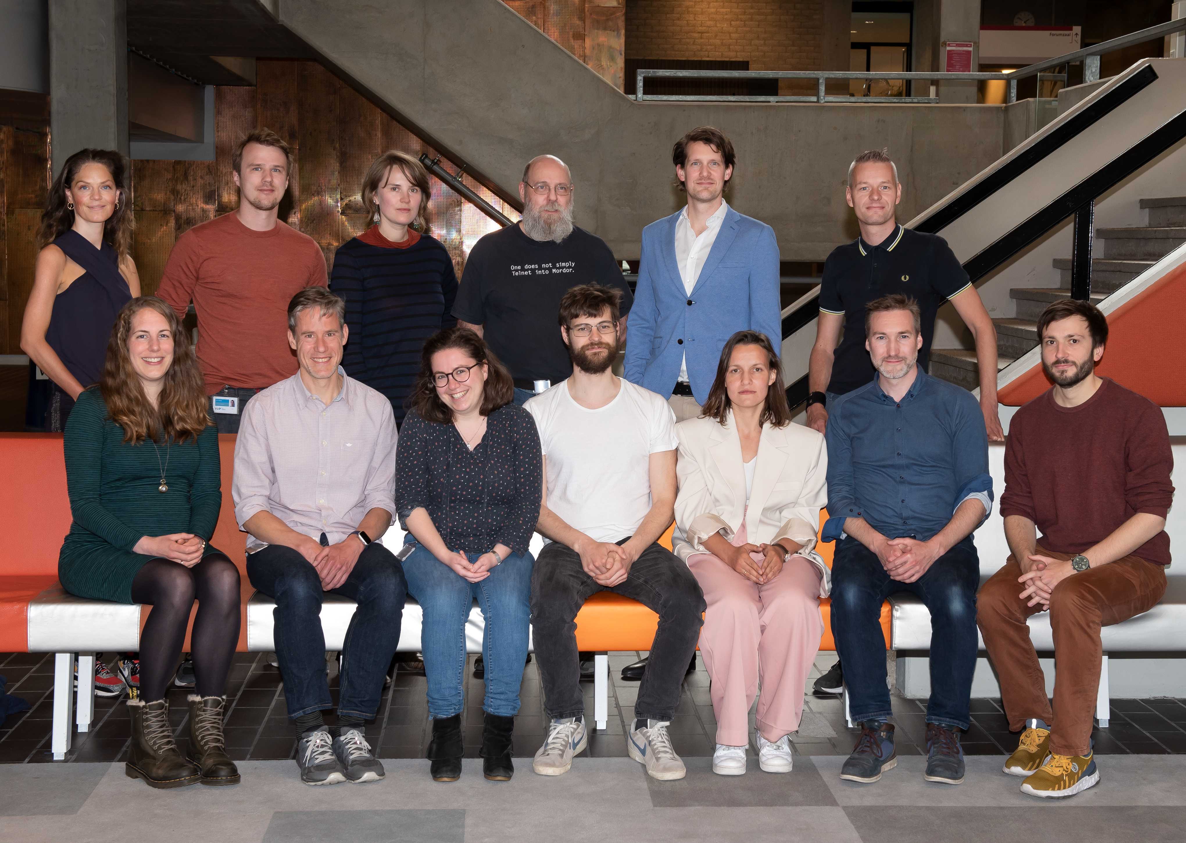 Thirteen people posing for a photo, half of them sitting and hald standing, like in a primary school photo. The background is a modern building with dark greys and oranges, with copper walls. Their ages seem the go from mid twenties to their fifties